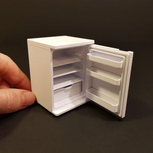 Puede incluir: Un refrigerador blanco en miniatura con una puerta que se abre para revelar estantes y un cajón en el interior. El refrigerador es aproximadamente del tamaño de la mano de una persona.