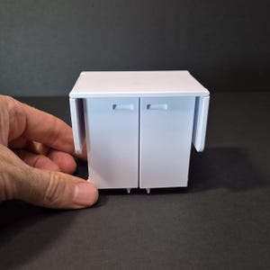 May include: A small, white cabinet with two doors and a fold-out side panel. The cabinet has a rectangular shape and is designed for a miniature setting. The doors have small, rectangular handles.