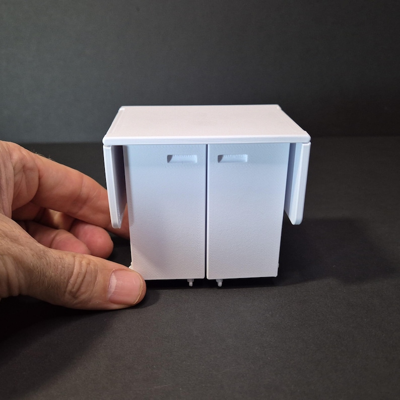 May include: A small, white cabinet with two doors and a fold-out side panel. The cabinet has a rectangular shape and is designed for a miniature setting. The doors have small, rectangular handles.