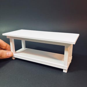 Peut inclure: Petite table rectangulaire blanche avec une étagère inférieure. La table mesure environ 30 cm de long, 15 cm de large et 10 cm de haut. Elle présente un design simple et moderne, faite d'un matériau opaque.