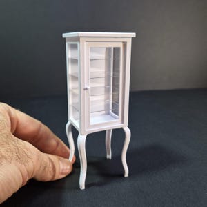 Op de afbeelding: Een witte miniatuur vitrinekast met glazen planken en een glazen deur. De kast heeft sierlijke, gebogen poten en een decoratieve rand rond de randen. De kast is ongeveer 15 cm hoog.