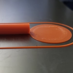 May include: Close-up of an orange 3D printed object with a circular design. The object is on a dark gray surface.