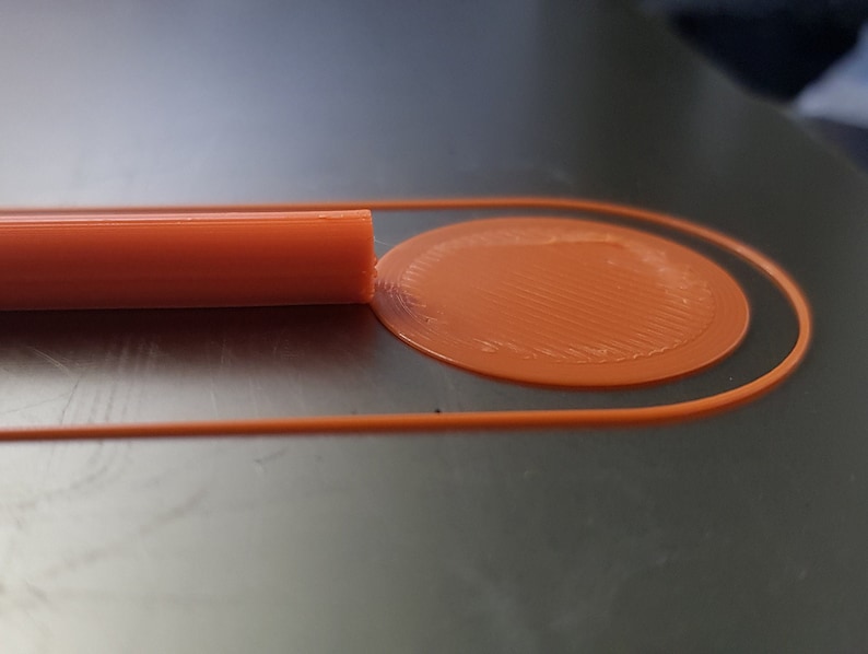 May include: Close-up of an orange 3D printed object with a circular design. The object is on a dark gray surface.