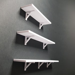 May include: Three white floating shelves with simple white brackets. The shelves are mounted on a dark gray wall.