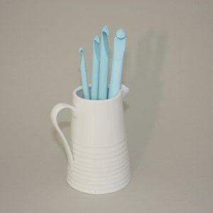May include: A white ceramic pitcher with a blue crochet hook set inside. The pitcher has a textured design and a handle.