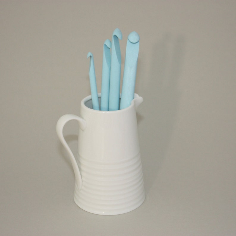 May include: A white ceramic pitcher with a blue crochet hook set inside. The pitcher has a textured design and a handle.