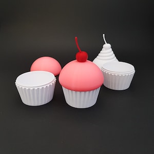 Puede incluir: Una colección de recipientes con forma de cupcake impresos en 3D. Un cupcake tiene una parte superior rosa con una cereza roja. Otros cupcakes son blancos y rosas. El fondo es negro.