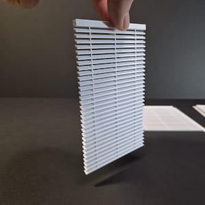 Puede incluir: Mini persiana veneciana blanca sostenida por una mano, proyectando una sombra sobre una superficie oscura. La persiana está compuesta por numerosas lamas horizontales. Se ven persianas adicionales en el fondo.