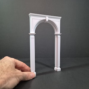 May include: A white architectural archway with a rounded top and two fluted columns. The archway is a miniature model, likely for a dollhouse or architectural project. The design features classical elements and is made of a solid, opaque material.