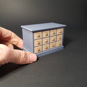 May include: A small, gray wooden dresser with 16 drawers. Each drawer has a gold knob and a light brown front.