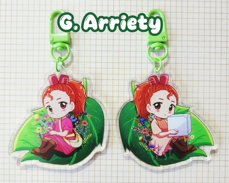 May include: Two keychains featuring a cartoon girl with red hair, sitting on a green leaf with flowers. The keychains have a green clasp and the text "G. Arrietty" in green.