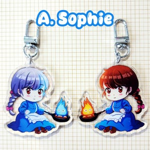 May include: Two acrylic keychains featuring anime-style illustrations of a character in a blue dress, holding a pan with a flame. One keychain has a character with purple hair, the other with red hair. The text "A. Sophie" is above.