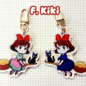 May include: Two acrylic keychains featuring a cartoon girl with a red bow, riding a broom with a black cat. One keychain has the girl in a pink and green dress, the other in a blue dress. The text "F. Kiki" is at the top.