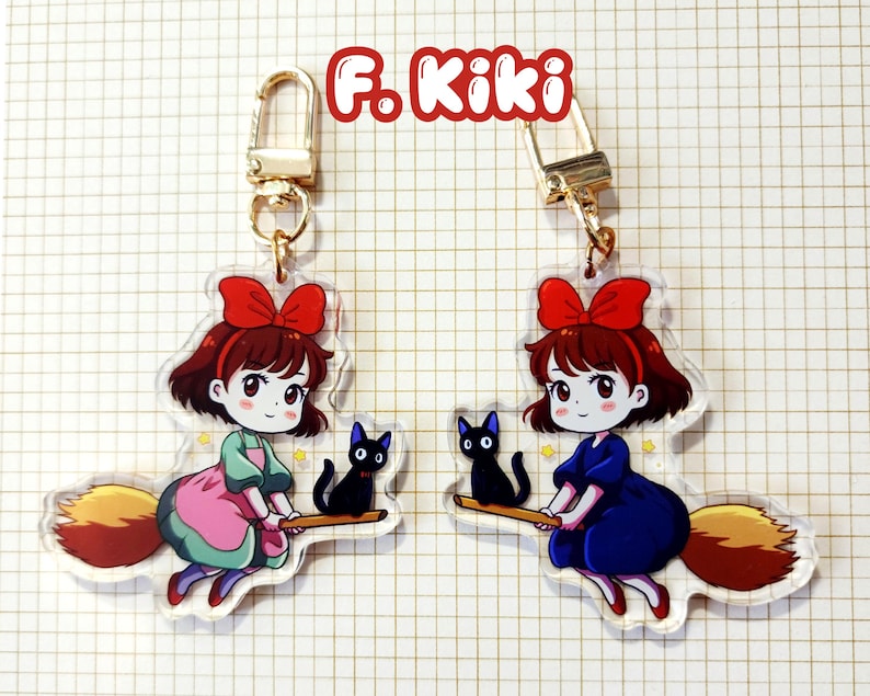 May include: Two acrylic keychains featuring a cartoon girl with a red bow, riding a broom with a black cat. One keychain has the girl in a pink and green dress, the other in a blue dress. The text "F. Kiki" is at the top.