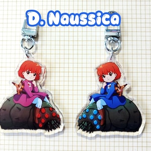 May include: Two acrylic keychains featuring a cartoon character with red hair and a small creature. The character is wearing a pink and blue outfit in one keychain and a blue outfit in the other. The keychains have silver-colored clasps and the text "D. Naussica".