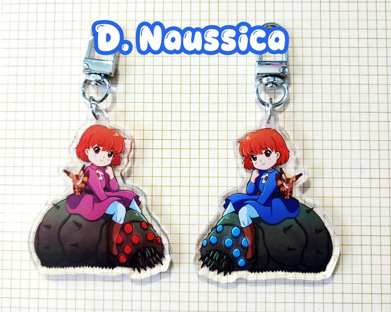 May include: Two acrylic keychains featuring a cartoon character with red hair and a small creature. The character is wearing a pink and blue outfit in one keychain and a blue outfit in the other. The keychains have silver-colored clasps and the text "D. Naussica".