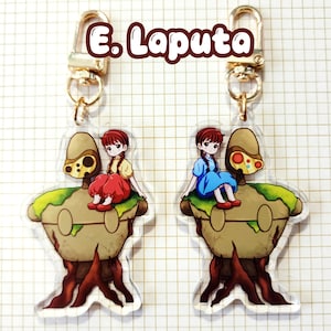 May include: Two acrylic keychains featuring a cartoon girl in a dress sitting on a brown and green structure with a tree trunk base. The keychains have gold-colored clasps and the text "E. Laputa" in a brown font.