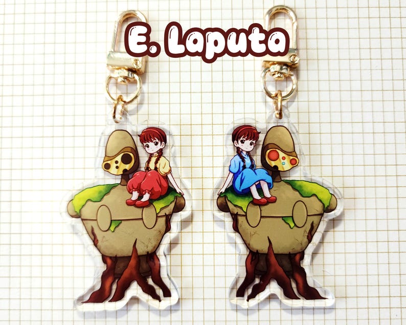 May include: Two acrylic keychains featuring a cartoon girl in a dress sitting on a brown and green structure with a tree trunk base. The keychains have gold-colored clasps and the text "E. Laputa" in a brown font.