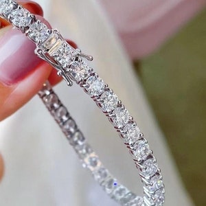 Może przedstawiać: Bransoletka tenisowa z rzędem okrągłych diamentów o szlifie brylantowym, osadzonych w srebrnej lub białego złota oprawie. Bransoletka jest zapinana na zapięcie. Diamenty błyszczą.