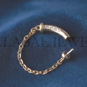 May include: A gold-colored earring with a curved bar set with small, clear stones. A delicate chain connects the bar to a post and clasp. The earring is displayed on a dark blue, textured surface. The text "GULABAE JEWELS" is visible in the background.