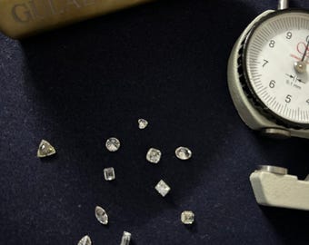 May include: A collection of small, sparkling diamonds in various shapes, including round, square, and pear-cut, are scattered on a dark blue surface. A gold-colored tool with the text "GULABAE JEWELS" is in the background, along with a measuring gauge.