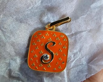 Puede incluir: Colgante naranja y dorado con la letra "S" en cursiva. El colgante tiene un borde dorado y una pequeña piedra con forma de diamante. El fondo naranja está decorado con formas de mariposas doradas.