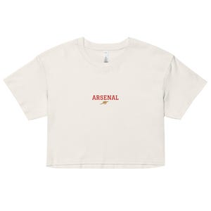 May include: A cream-coloured crop top with short sleeves. The word "ARSENAL" is embroidered in red, with a small emblem beneath it. The shirt is made of a soft material, suitable for casual wear.