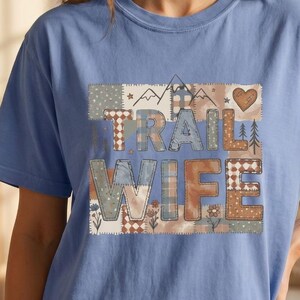 May include: A blue short-sleeve t-shirt featuring a rectangular graphic with the text TRAIL WIFE. The design consists of a patchwork pattern in shades of brown, blue, and cream, decorated with small illustrations of mountains, trees, and hearts.
