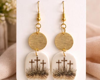 Puede incluir: Pendientes colgantes dorados. Cada pendiente presenta un disco dorado circular sobre un colgante ovalado. El colgante muestra tres cruces sobre una colina cubierta de hierba. La tarjeta dice "OLIVA COLLECTIVE handcrafted jewelry."