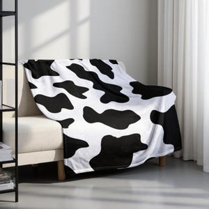 May include: A white blanket with large black cow spots draped over a beige sofa. The blanket is made of a soft material and adds a decorative touch to the room. The blanket is a cozy home decor item.