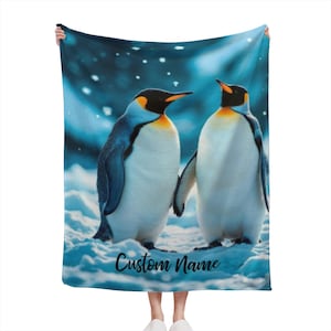 May include: A blue blanket featuring two penguins in a snowy landscape. The penguins have black heads, yellow accents, and white bodies. The blanket has the text "Custom Name" in a script font.
