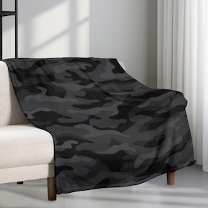 May include: A dark grey camouflage pattern blanket draped over a beige sofa. The blanket features a repeating design of irregular shapes in varying shades of grey and black, creating a military-inspired aesthetic. The blanket is soft and cosy.