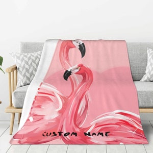 May include: Pink fleece throw blanket featuring a watercolour illustration of two pink flamingos with black eyes and beaks. The text "CUSTOM NAME" is written in black at the bottom of the blanket.