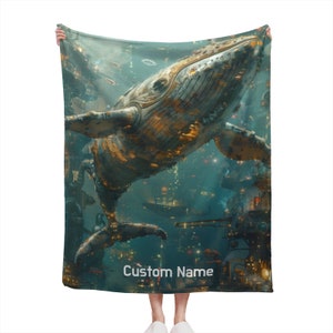 Può includere: Una coperta in pile blu turchese con una grande illustrazione dettagliata di una balena in stile steampunk. La balena è fatta di metallo e ha luci e ingranaggi. Il testo "Custom Name" è stampato sulla coperta.