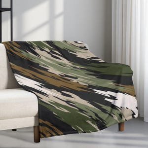 May include: A camouflage-style blanket in shades of green, brown, black, and white, draped over a beige sofa. The blanket's design consists of diagonal brushstrokes. The sofa has wooden legs and is near a window with white curtains.