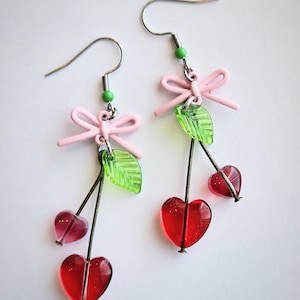 May include: A pair of dangle earrings featuring red heart-shaped cherries, green leaves, and pink bows. The earrings have silver-colored hooks and stems. The cherries are made of a translucent material.