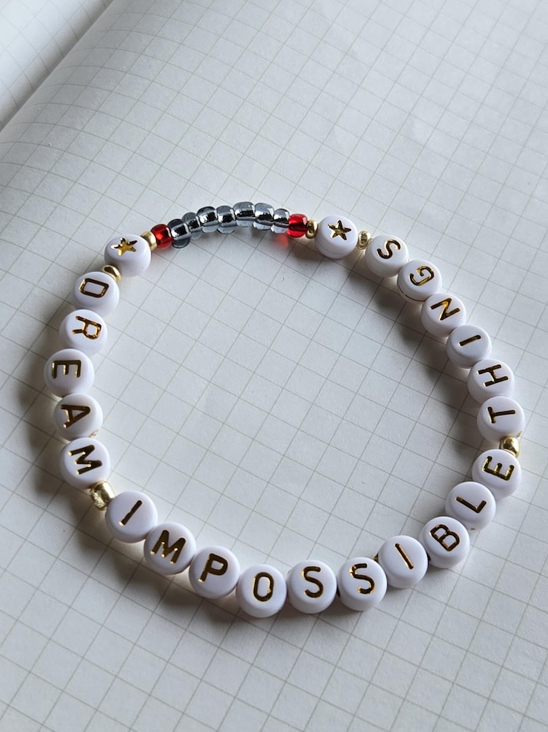 May include: White beaded bracelet with gold lettering spelling "DREAM IMPOSSIBLE". The bracelet includes gold star beads, red beads, and silver beads. The bracelet is on a white grid background.