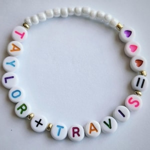 May include: A white beaded bracelet with the names "Taylor + Travis" spelled out in colorful letter beads. The bracelet also features heart-shaped beads and gold-colored accents.