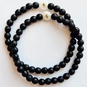 May include: Two beaded bracelets. Each bracelet is made of black, round beads. Each bracelet has a white bead with a gold lightning bolt design.