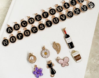 May include: A set of gold-toned charms featuring letters of the alphabet, a wine bottle, a coffee cup, a seashell, a flower, a butterfly, a leaf, and a cat. The charms are black and white with gold accents.