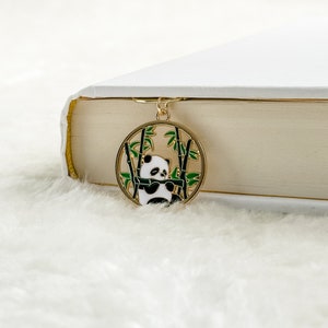 Puede incluir: Marcapáginas de metal dorado con un panda sentado en un bosque de bambú. El panda es blanco y negro, y el bambú es verde.