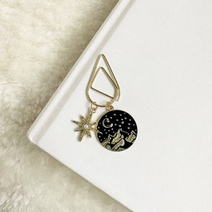 May include: Gold bookmark with a black circle charm featuring a crescent moon, stars, and mountains. The charm is attached to a gold clip with a star charm.