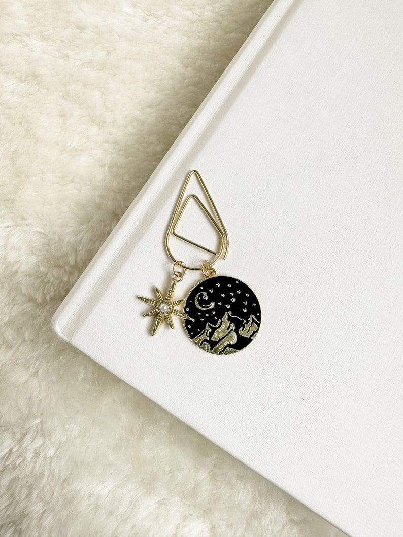 May include: Gold bookmark with a black circle charm featuring a crescent moon, stars, and mountains. The charm is attached to a gold clip with a star charm.