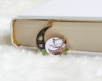 May include: Gold metal bookmark featuring a white cat curled up on a black crescent moon with pink and green floral accents.