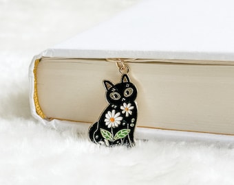 May include: A black cat bookmark with white flowers and green leaves. The bookmark is made of metal and has a gold finish.