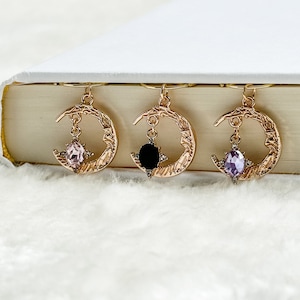 May include: Three gold crescent moon bookmark charms with different coloured gemstones. The charms are hanging from a book.