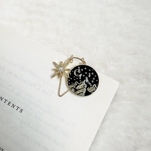 May include: Gold bookmark with a black circle featuring a crescent moon, stars, and mountains. A gold star with a pearl in the centre is attached to the circle.