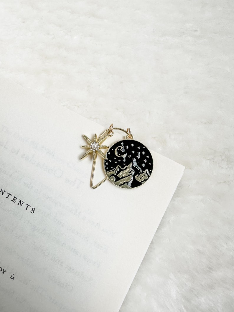 May include: Gold bookmark with a black circle featuring a crescent moon, stars, and mountains. A gold star with a pearl in the centre is attached to the circle.