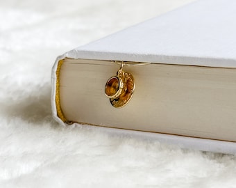 Może przedstawiać: Złoty zakładka w kształcie filiżanki do herbaty i spodeczka przymocowana do białej książki.