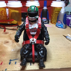 Puede incluir: Un asiento de carreras negro con arnés de carreras rojo y un casco verde y blanco. El asiento está etiquetado como "CORE AUTO" y el casco tiene la palabra "SIMPSON" en él. El casco también tiene las palabras "INTERSTATE BATTERIES" en él.
